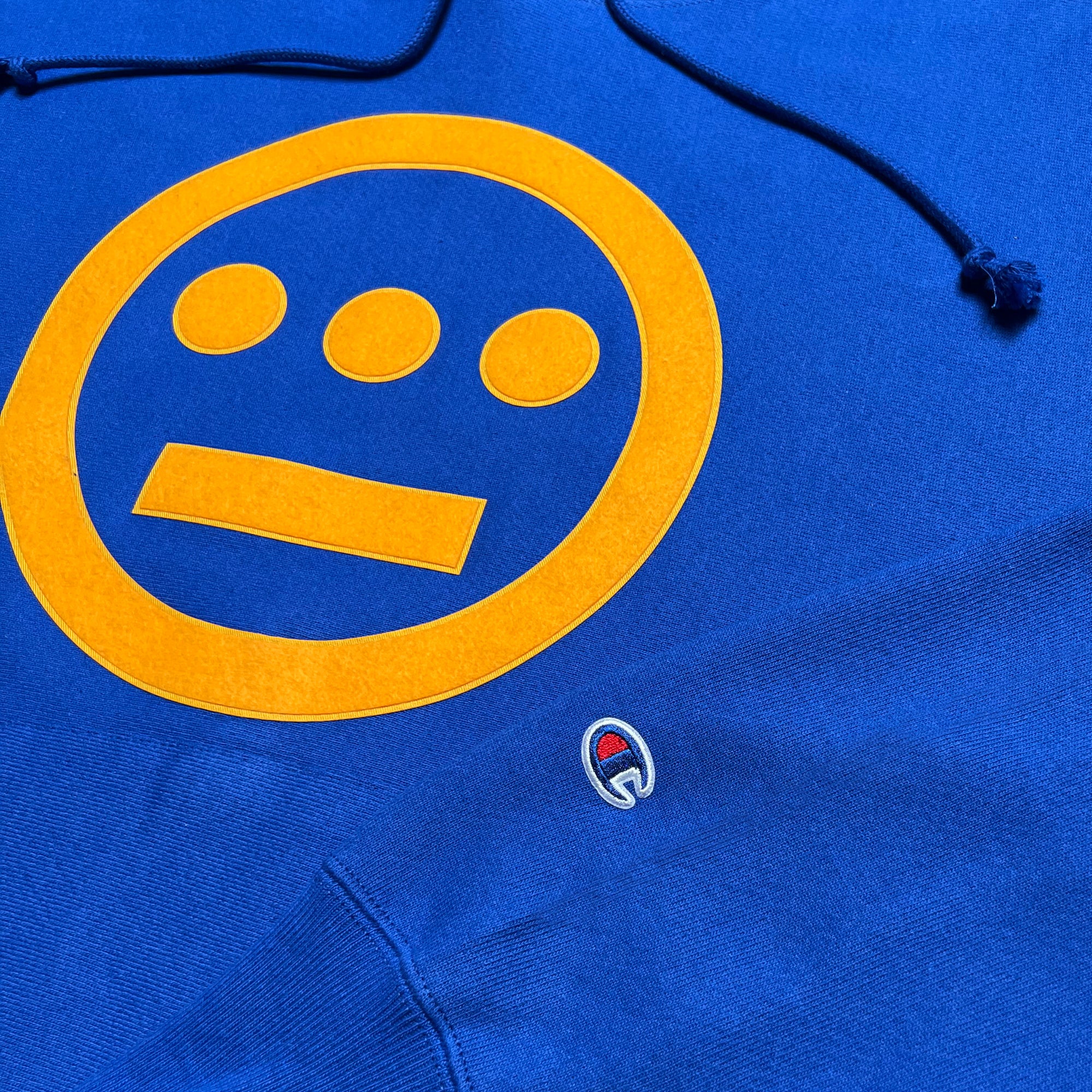 Close-up of the Champion X Hiero 3rd Eye Hoodie, a blue heavyweight cotton fleece creation by Hieroglyphics, showcasing a large yellow logo with a circular face and three dots for eyes above a horizontal line for a mouth. An embroidered "C" in red, white, and blue beneath it signifies the Champion Reverse Weave brand.