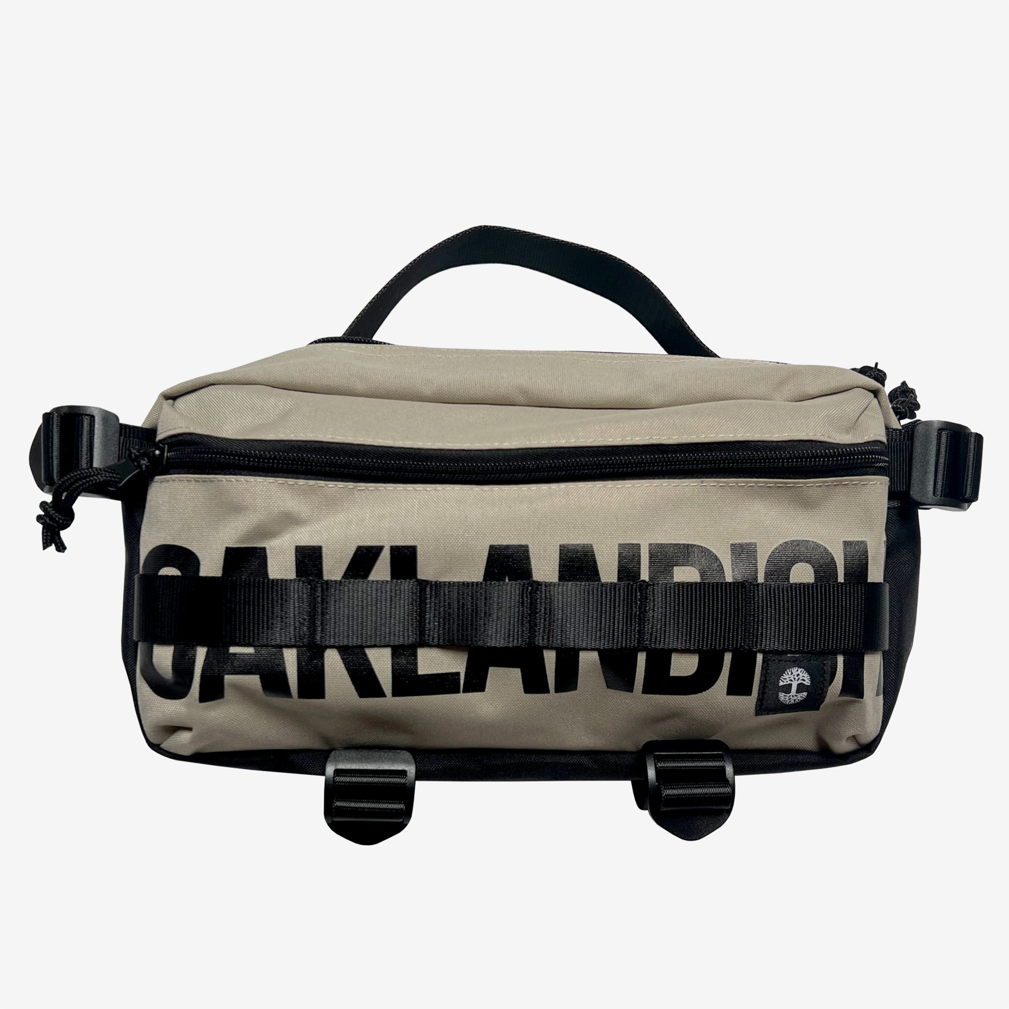 A sand brown hip bag with a black Oaklandish wordmark, top zipper & handle, waist belt, & small white Oaklandish tree logo.