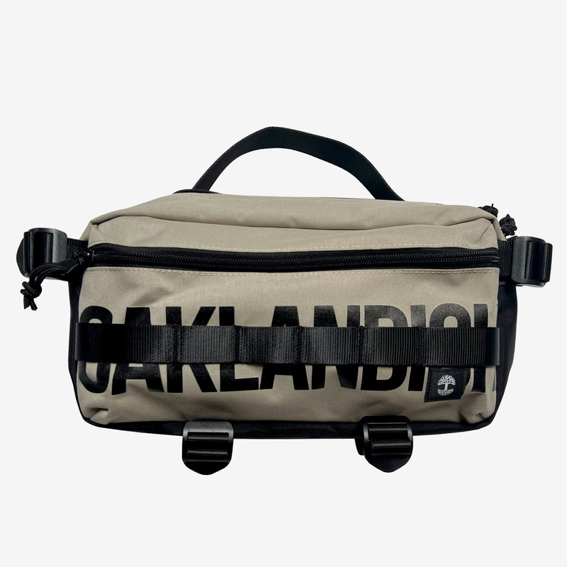 A sand brown hip bag with a black Oaklandish wordmark, top zipper & handle, waist belt, & small white Oaklandish tree logo.