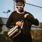 Male model outside wearing tan nylon hip bag with a black Oaklandish wordmark & small white Oaklandish tree logo.
