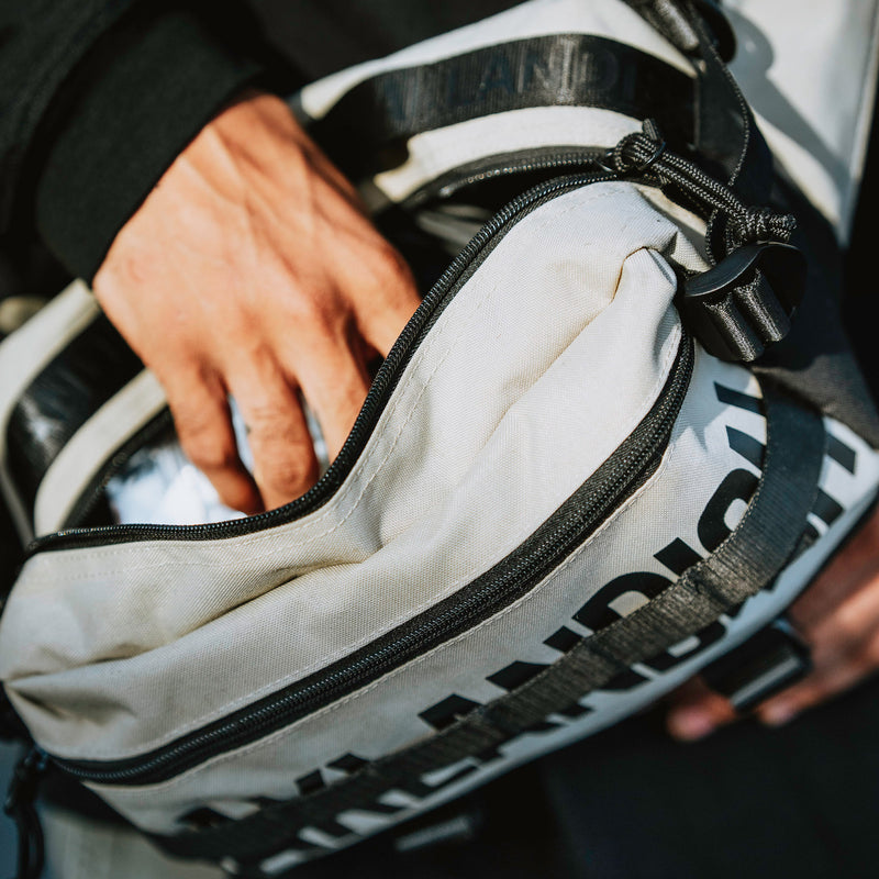 Detailed image of hand reaching into a cross body tan hip bag with a Oaklandish wordmark and tree logo.
