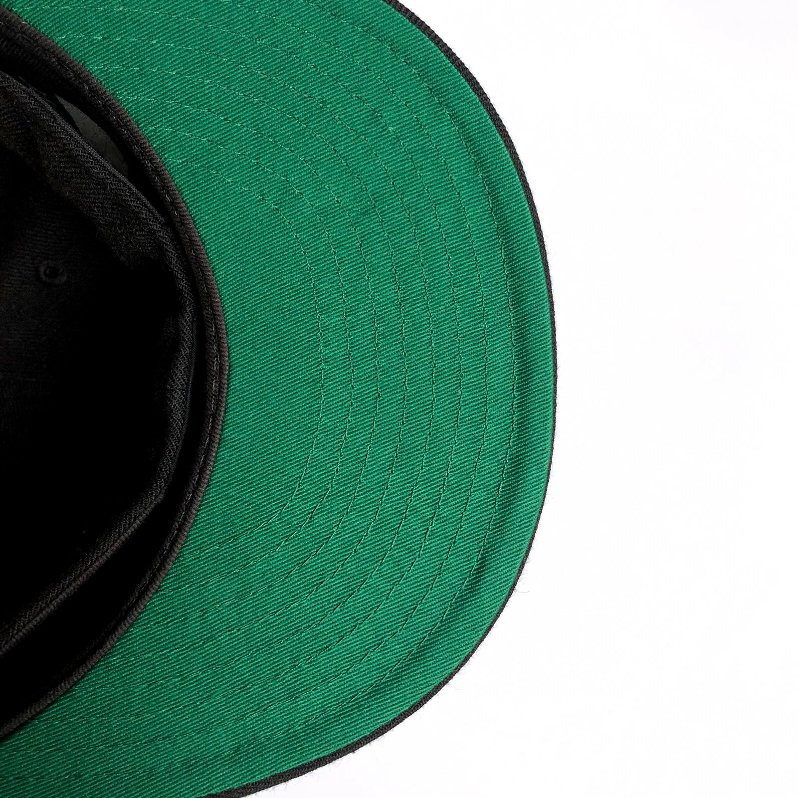 Close-up of the underside of an Oaklandish New Era OAK 59FIFTY Fitted Cap. The brim's underside is green, while the inner part of the cap is black. The right portion of the image shows a white background, providing contrast against the cap's colors. The stitching and texture of the fabric are clearly visible.