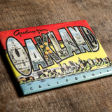 Full color, red, blue, yellow with black lettering, “Greetings for Oakland, California” postcard style magnet on wood background.