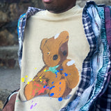 Man’s chest in a brown t-shirt with an artistically painted battered teddy bear with yellow and red paint splatter.