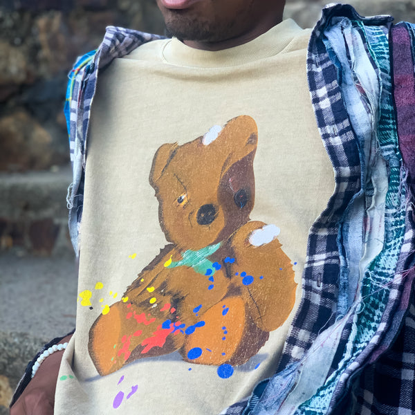 Man’s chest in a brown t-shirt with an artistically painted battered teddy bear with yellow and red paint splatter.