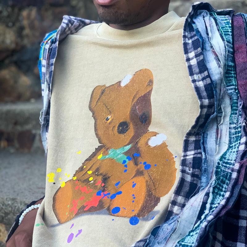 Man’s chest in a brown t-shirt with an artistically painted battered teddy bear with yellow and red paint splatter.