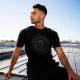 A man sitting on a dock on the water wearing a black t-shirt with a full circle black Roots SC logo, looking sideways.