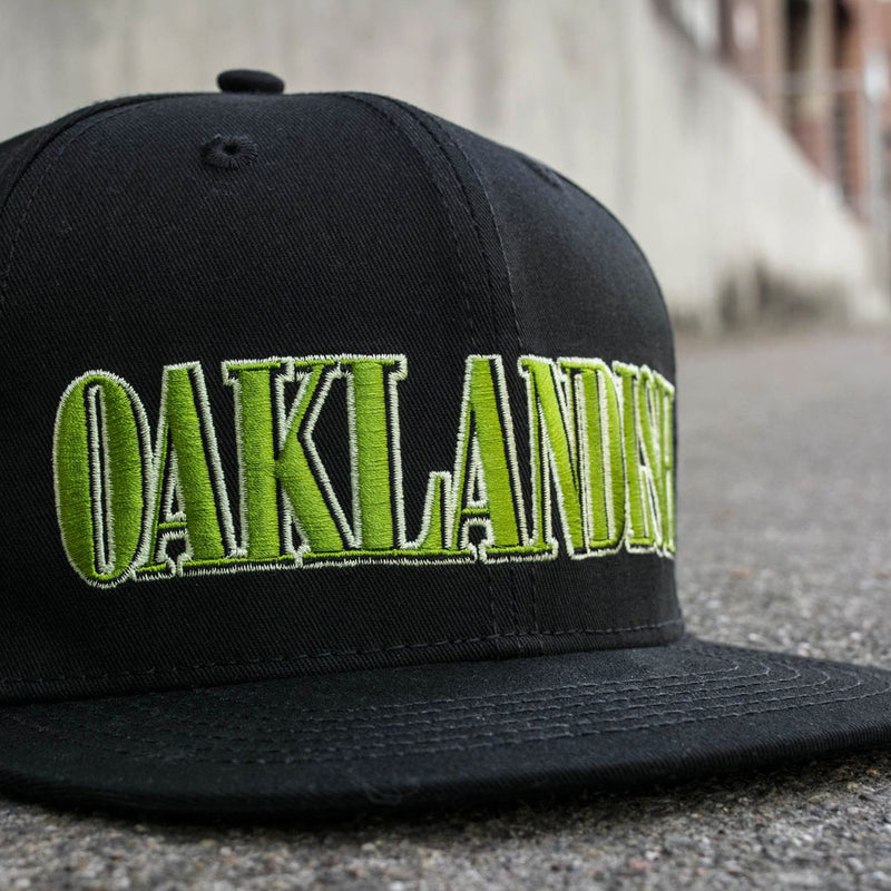 Side view of a black snapback cap with green embroidered Oaklandish tree logo on the front outdoors on the sidewalk. 