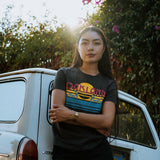Female model outside in a faded black women’s t-shirt with multi-color license plate design with capital letters spelling out OAKLAND.