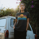 Female model outside in a faded black women’s t-shirt with multi-color license plate design with capital letters spelling out OAKLAND.
