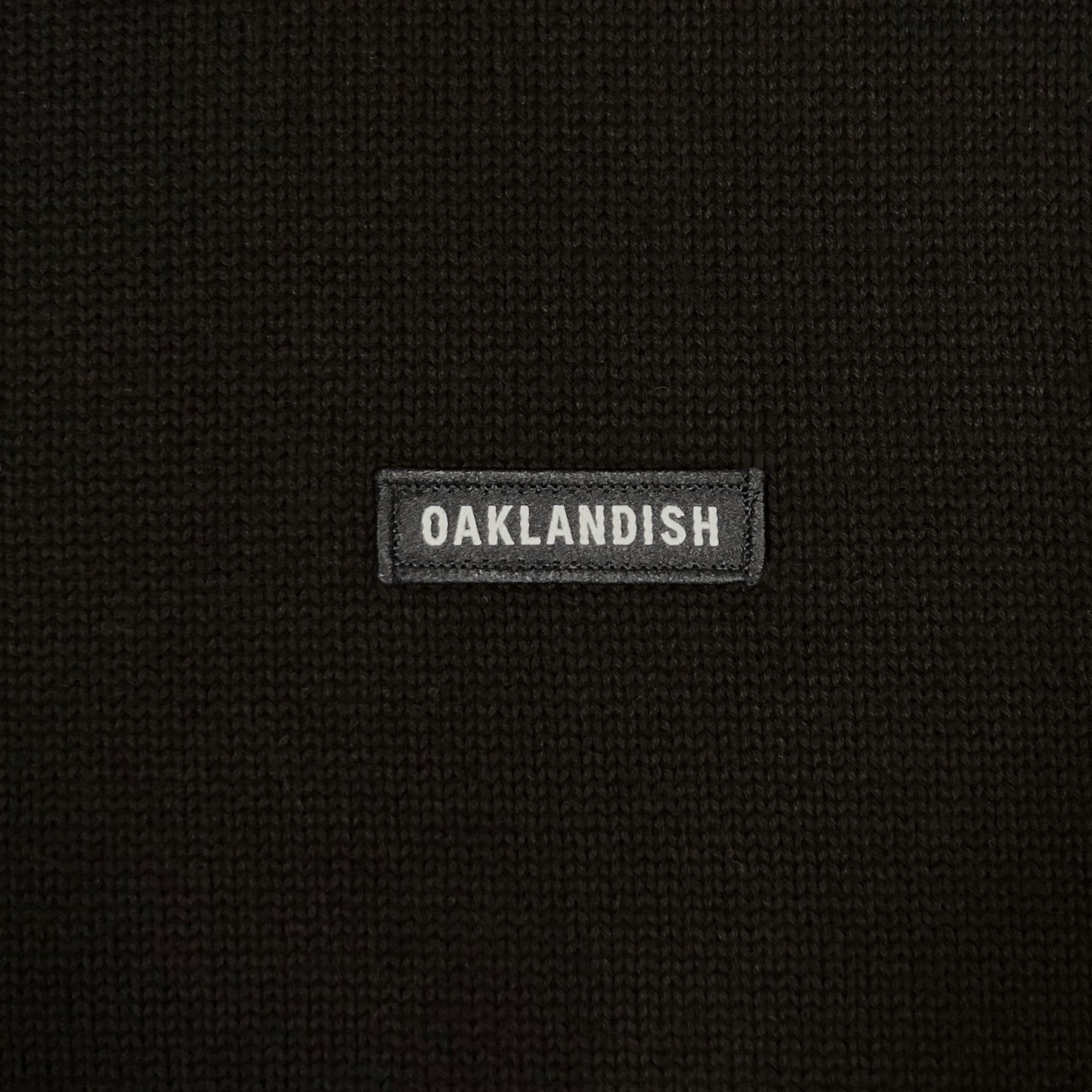 Detailed close-up of embroidered Oaklandish wordmark woven label patch on a black heavy-knit sweater.