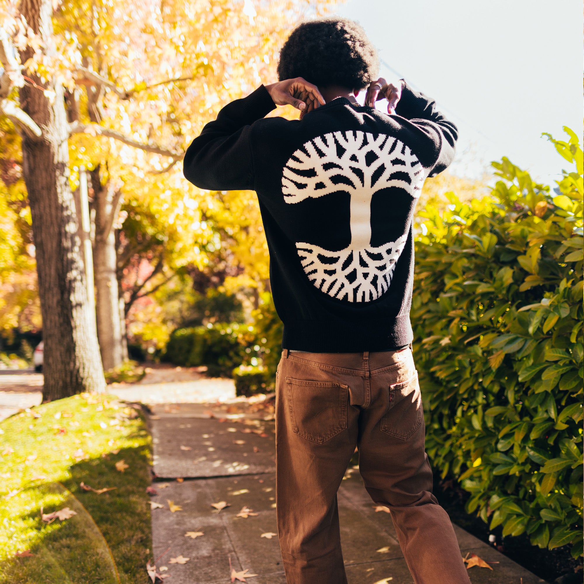 A man walking outside in a heavy-weight knit, black sweater with a large white Oaklandish tree logo on the back.