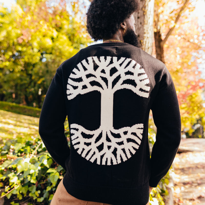 A man standing outside in a heavy-weight knit, black sweater with a large white Oaklandish tree logo on the back.