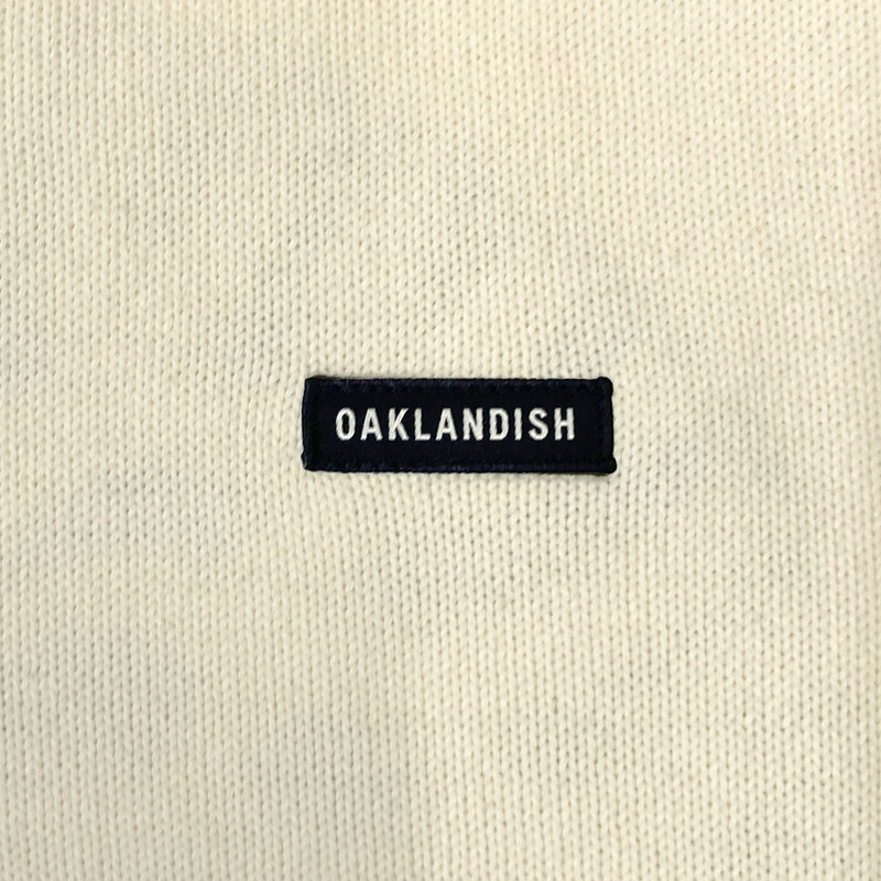 Detailed close-up of embroidered Oaklandish wordmark woven label patch on a white heavy-knit sweater.