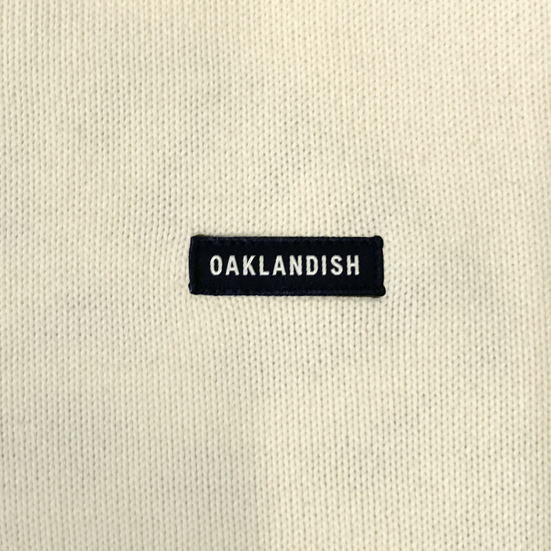 Detailed close-up of embroidered Oaklandish wordmark woven label patch on a white heavy-knit sweater.