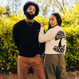 A man and a woman are standing outdoors in heavy-knit sweaters, the man facing forward in black and the woman facing backward in white.