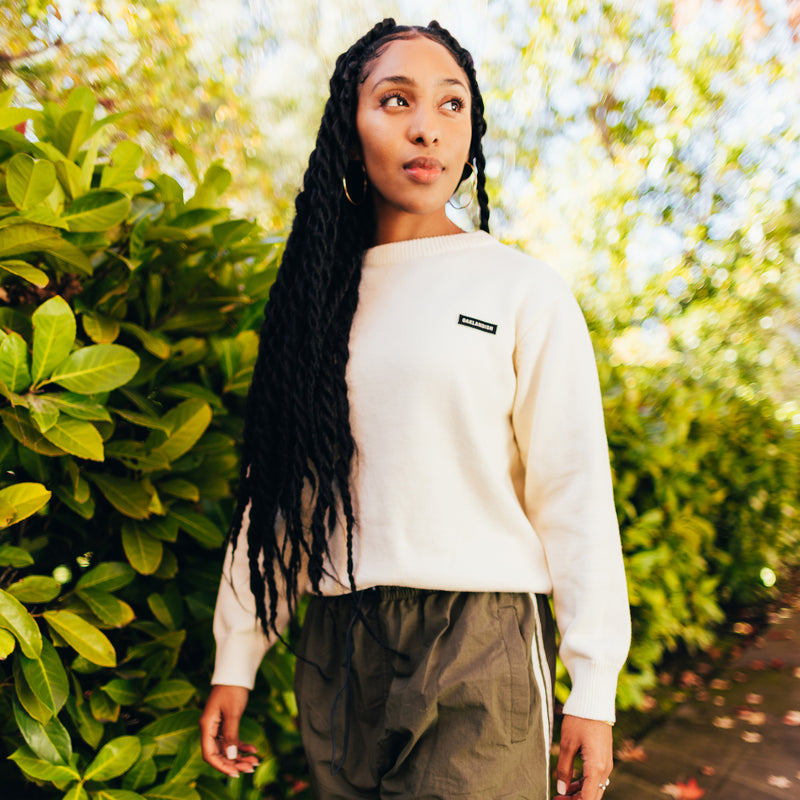 A woman outside, from the waist up, in a white knit sweater with embroidered Oaklandish wordmark label on the front left chest.
