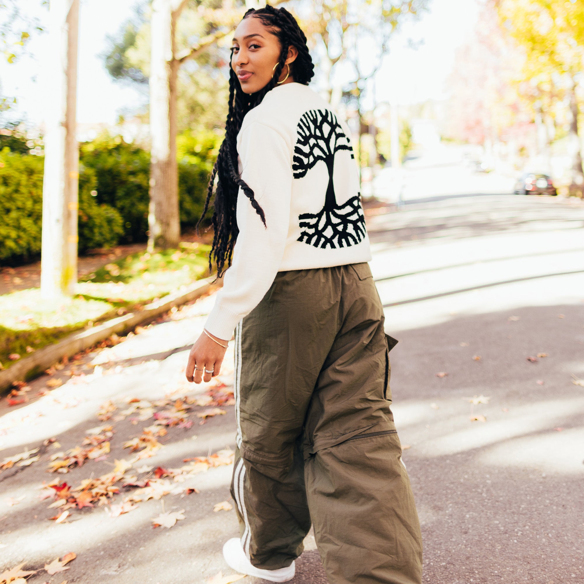 A woman walking outside in a white knit sweater with a large black Oaklandish logo on the back.