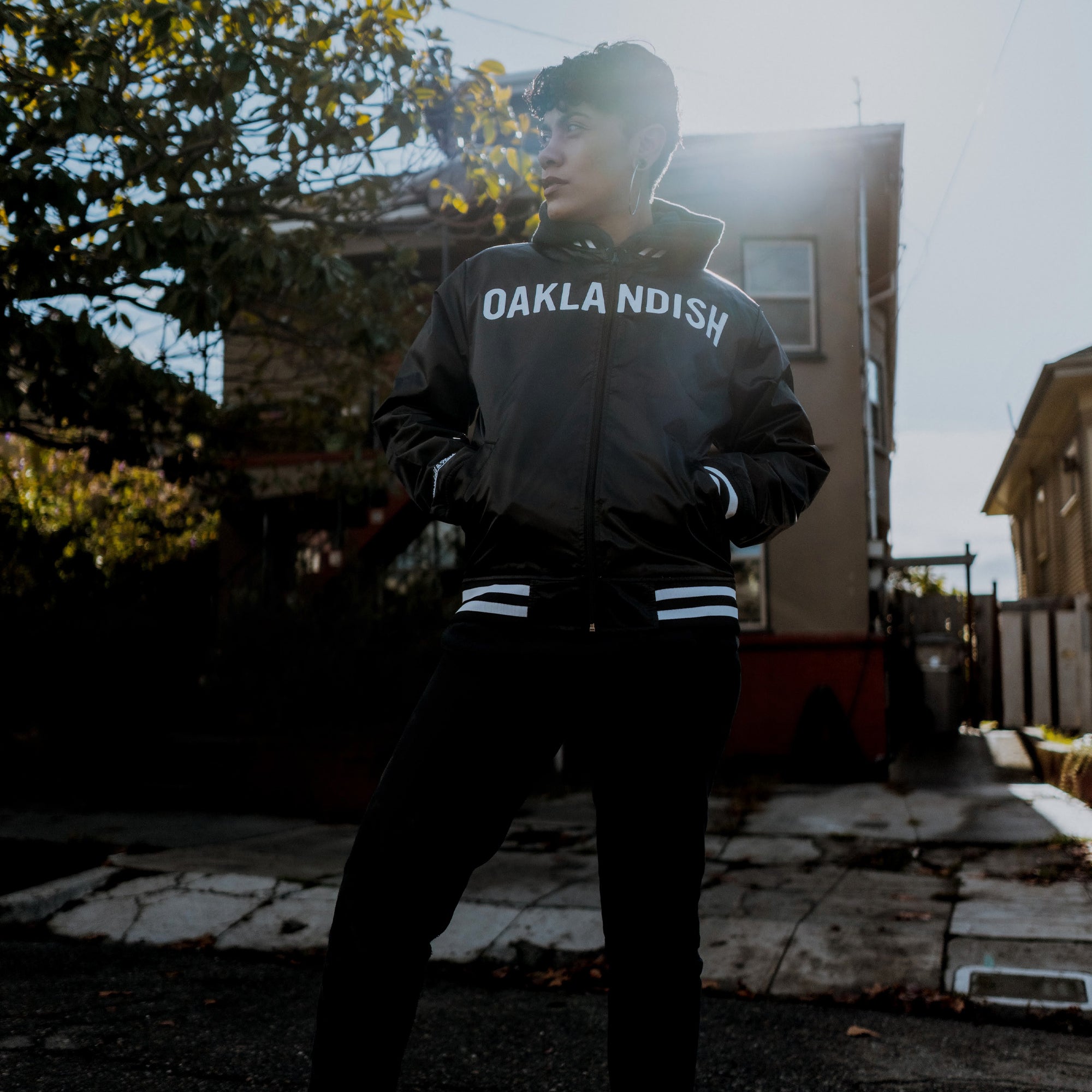An individual wearing a Mitchell & Ness x Oaklandish Reversible Satin Jacket stands in a residential area. With short hair, they gaze sideways as sunlight creates dramatic backlighting. A house and tree are seen in the background, with sunlight patches on the ground.