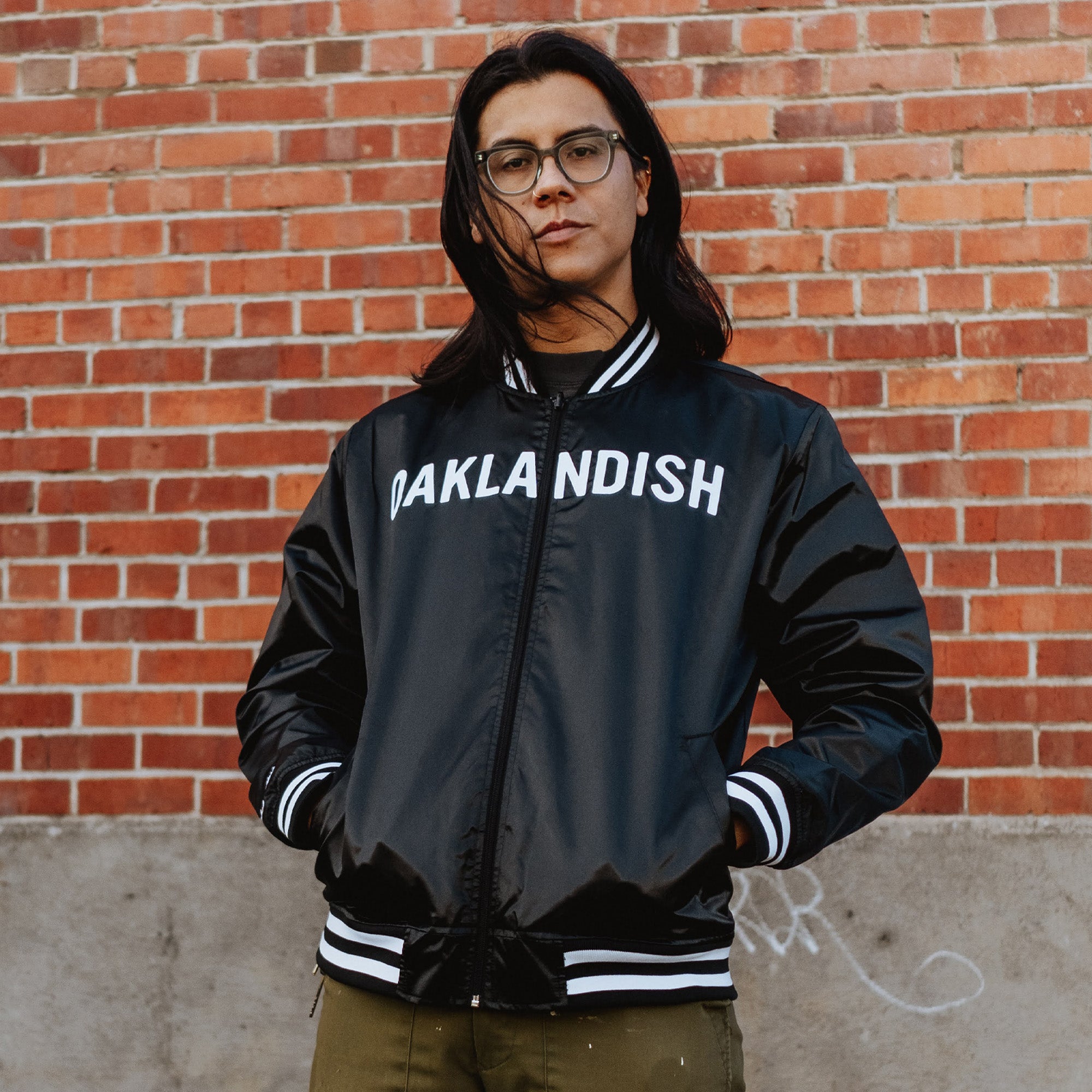 A confident person with long black hair and glasses stands in front of a brick wall wearing the Mitchell & Ness x Oaklandish Reversible Satin Jacket, featuring "OAKLANDISH" in white. Olive-green pants accentuate the jacket's striped cuffs and collar as they gaze directly at the camera.