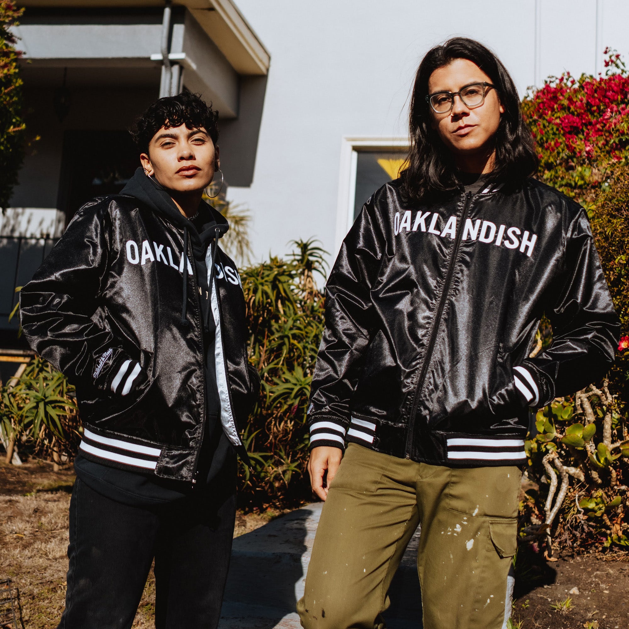 Two people stand outside a building, both wearing black Mitchell & Ness x Oaklandish Reversible Satin Jackets. The person on the left, with short curly hair and the hood up, is next to someone with long straight hair and glasses. They are framed by a garden, highlighting Oaklandish style beautifully.