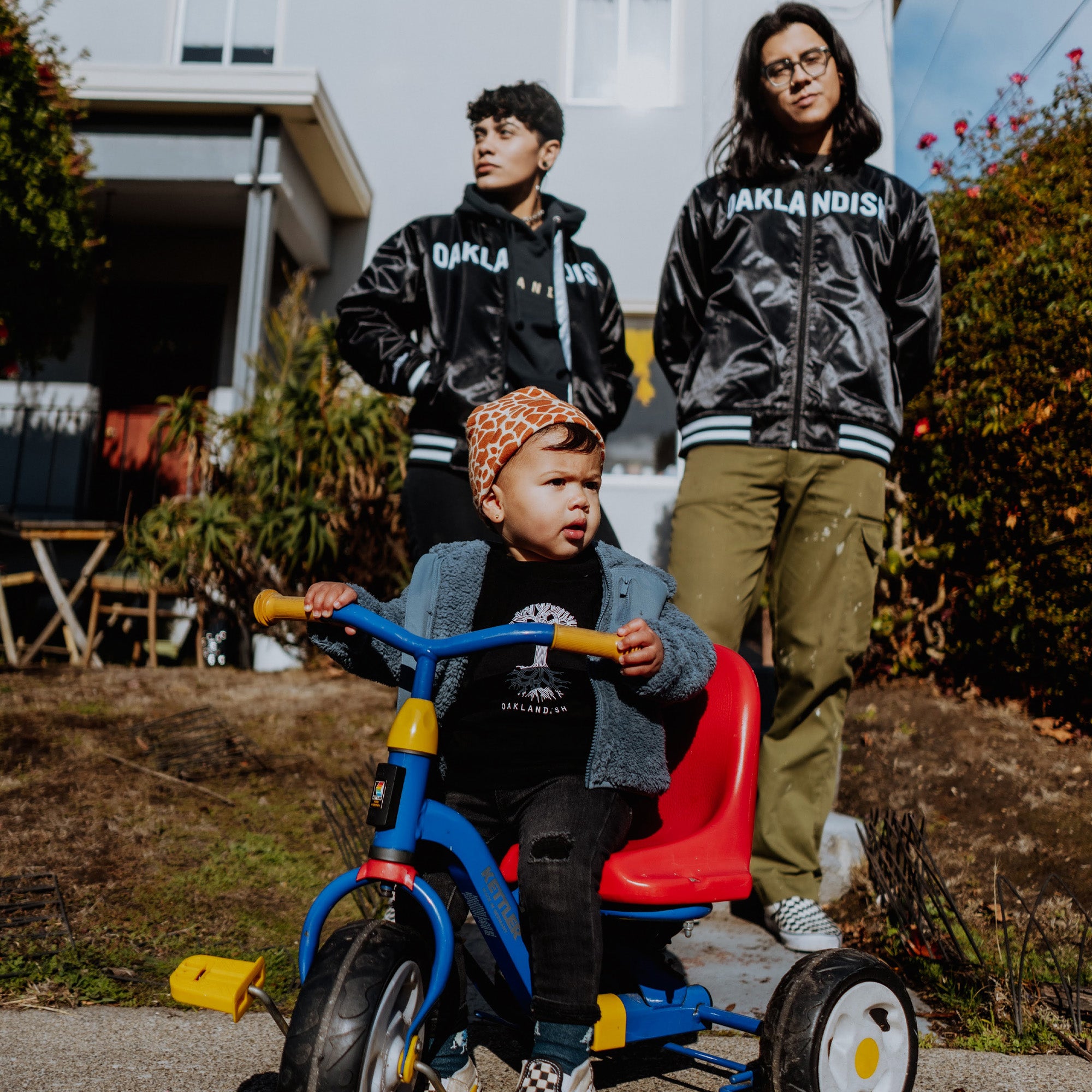 A toddler in a patterned hat sits on a colorful tricycle. Behind them, two adults wearing matching Mitchell & Ness x Oaklandish reversible satin jackets stand on a sunny sidewalk; one has curly hair, the other wears glasses. A house and garden appear in the background.