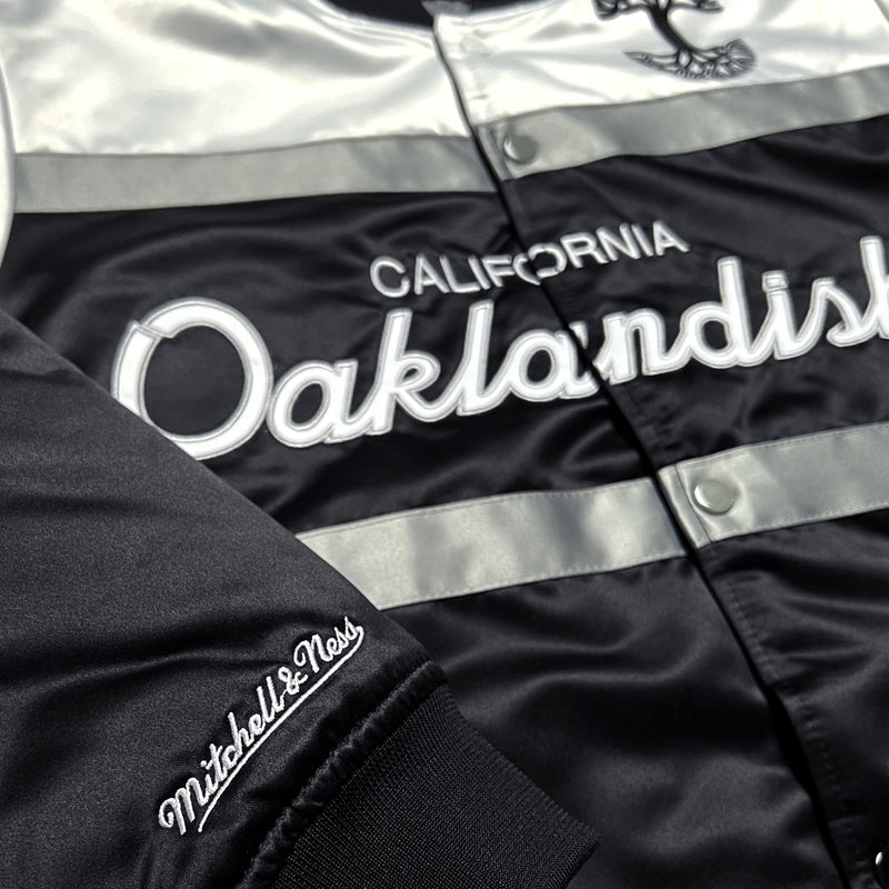 Close-up of Mitchell & Ness wordmark on the sleeve and California Oaklandish wordmark on the chest of a striped satin jacket.