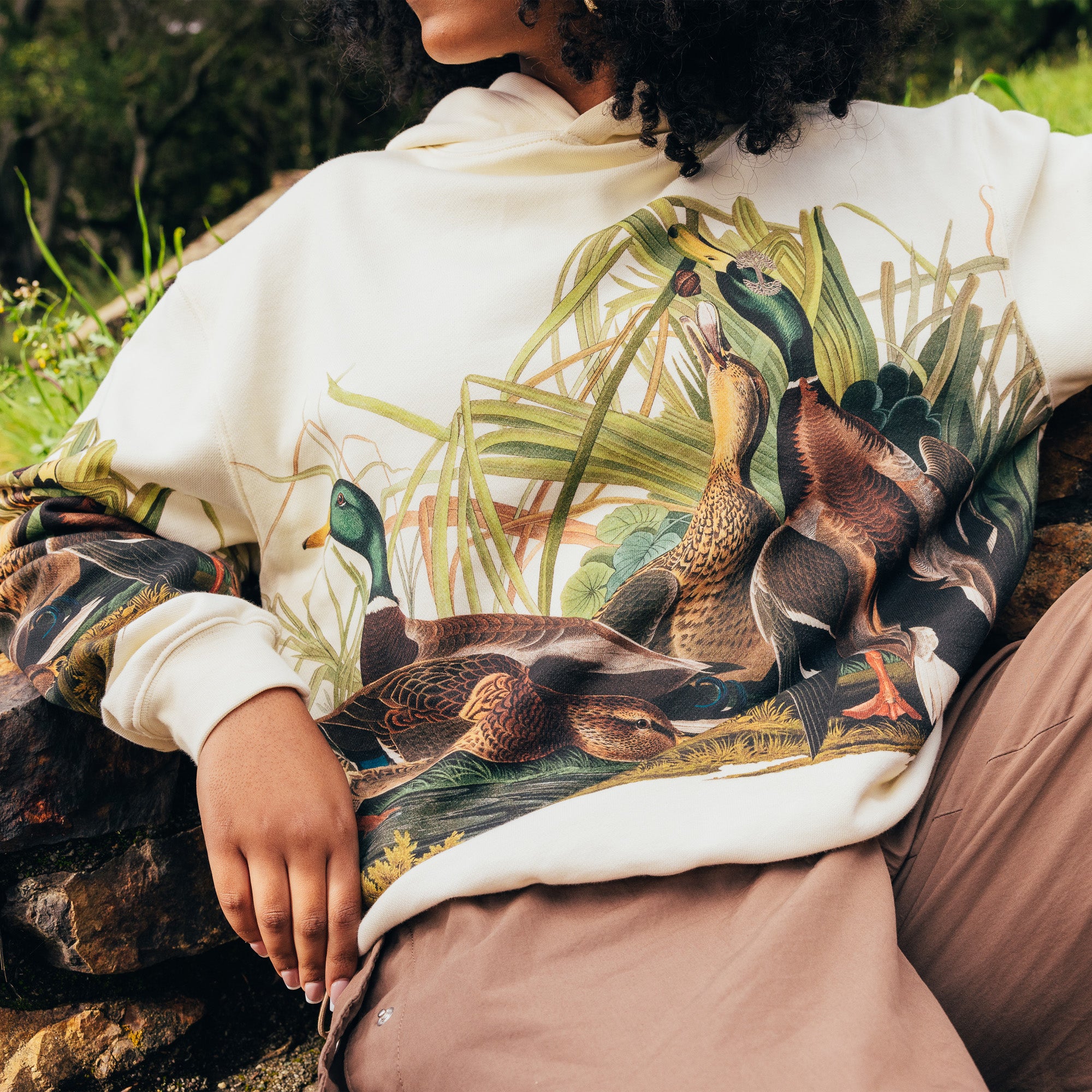 Close-up of the chest of a woman sitting outdoors wearing a cream pullover Oaklandish hoodie with Mallards printed all over. 