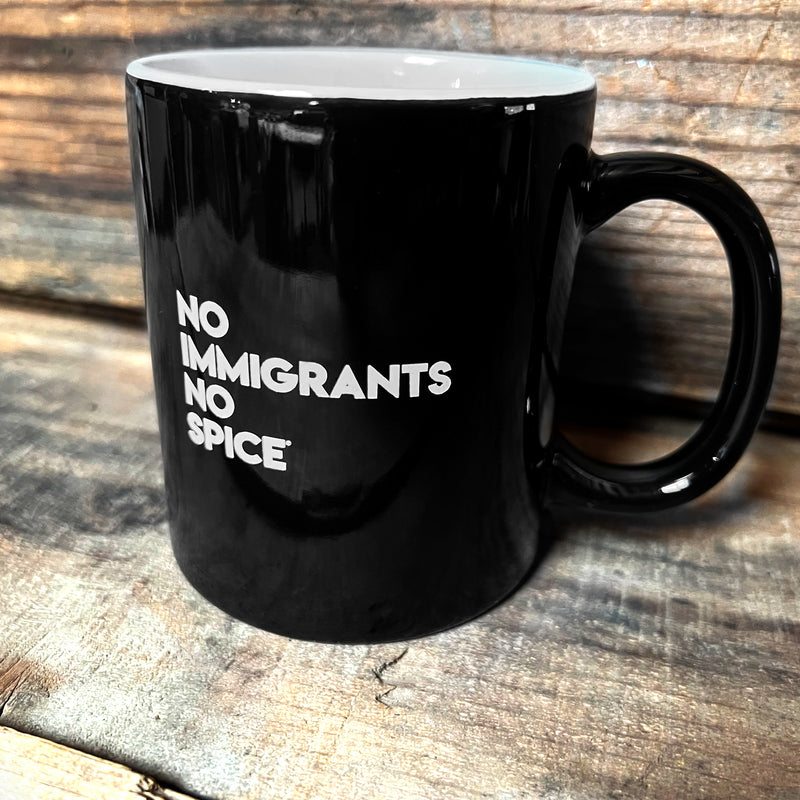 Glossy finish black ceramic coffee mug with white NO IMMIGRANTS NO SPICE wordmark on wood shelf.