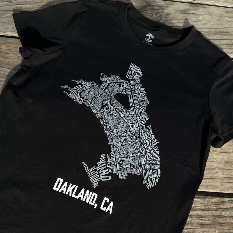 Black t-shirt with an Oakland neighborhood map with Oakland CA wordmark on a wood deck.