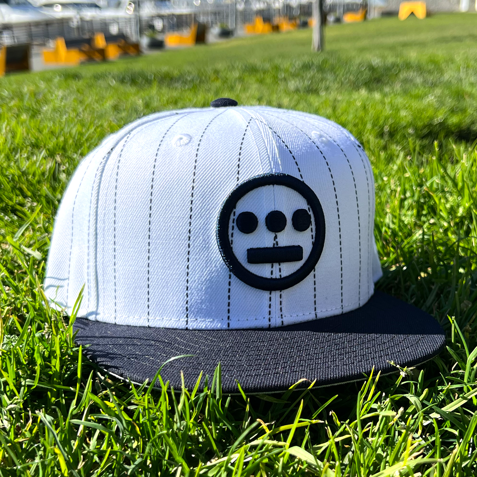 A New Era Hiero Pinstripe 59FIFTY Fitted Cap, featuring a white base with black pinstripes and a flat black bill, rests on vibrant green grass. The front of the hat showcases a black embroidered Hieroglyphics logo, characterized by a horizontal line with three dots above and two dots below. In the background, boats are docked at a marina.
