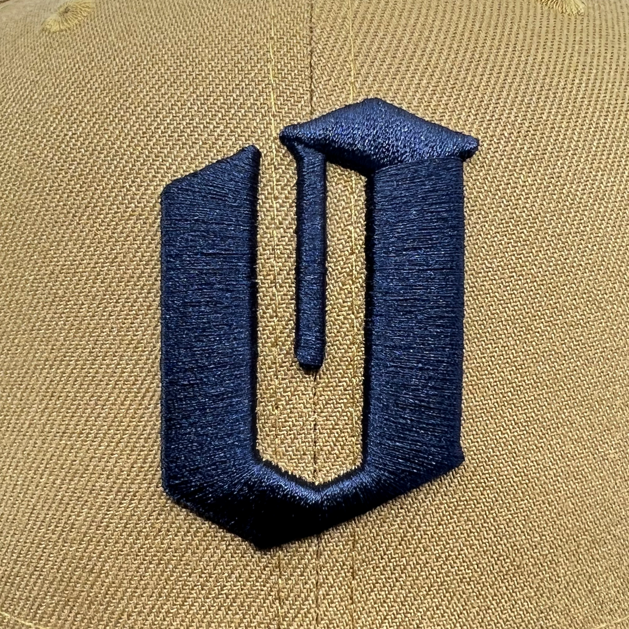 A close-up image of the front part of a beige New Era Official 59FIFTY Fitted Cap by Oaklandish, featuring a black, stylized letter "U" embroidered on it. The letter "U" boasts a modern, geometric design with its right vertical line extending downward past the baseline of the letter, creating a unique, elongated effect.