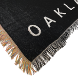 Detailed close-up of the corner of an Oaklandish throw blanket with multi-color fringe on one side and black and white fringe on the other.