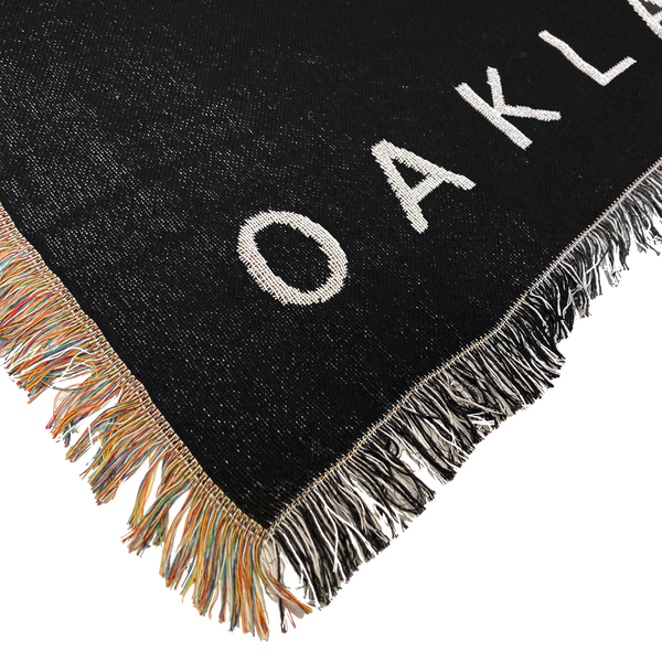 Detailed close-up of the corner of an Oaklandish throw blanket with multi-color fringe on one side and black and white fringe on the other.