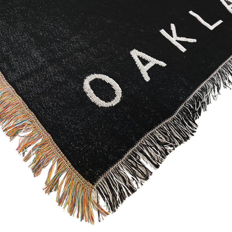 Detailed close-up of the corner of an Oaklandish throw blanket with multi-color fringe on one side and black and white fringe on the other.