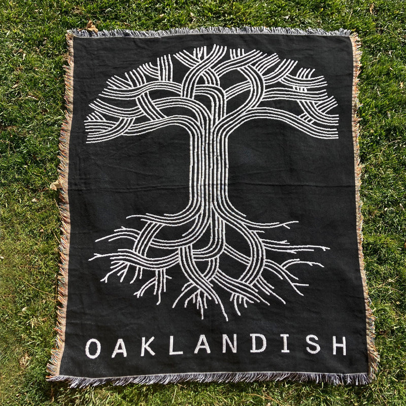 Black throw blanket with a large white Oaklandish tree logo and wordmark with fringe laying on the grass.