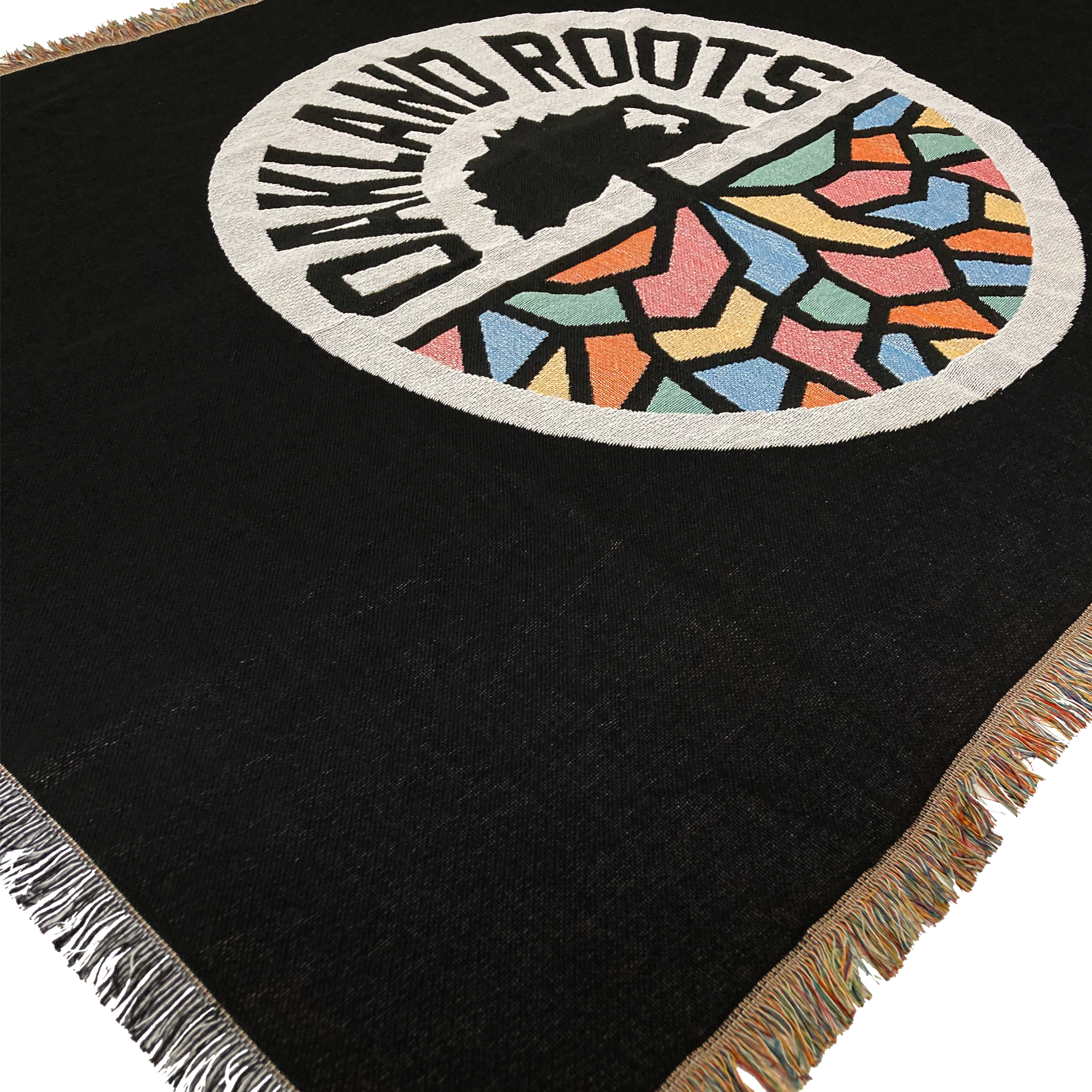 The Oakland Roots SC Throw by Oakland Roots SC is a woven black blanket adorned with a circular emblem. The center of the emblem showcases a tree, with "Oakland Roots Sports Club" arched above it. Below the tree are colorful geometric shapes in blue, red, orange, yellow, and green. The blanket is completed with multicolored fringe edges and radiates social good vibes.