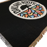 Detailed close-up of a black blanket with colored fringe and large full-color Oakland Roots SC circle logo in the center.