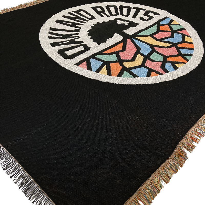 Detailed close-up of a black blanket with colored fringe and large full-color Oakland Roots SC circle logo in the center.