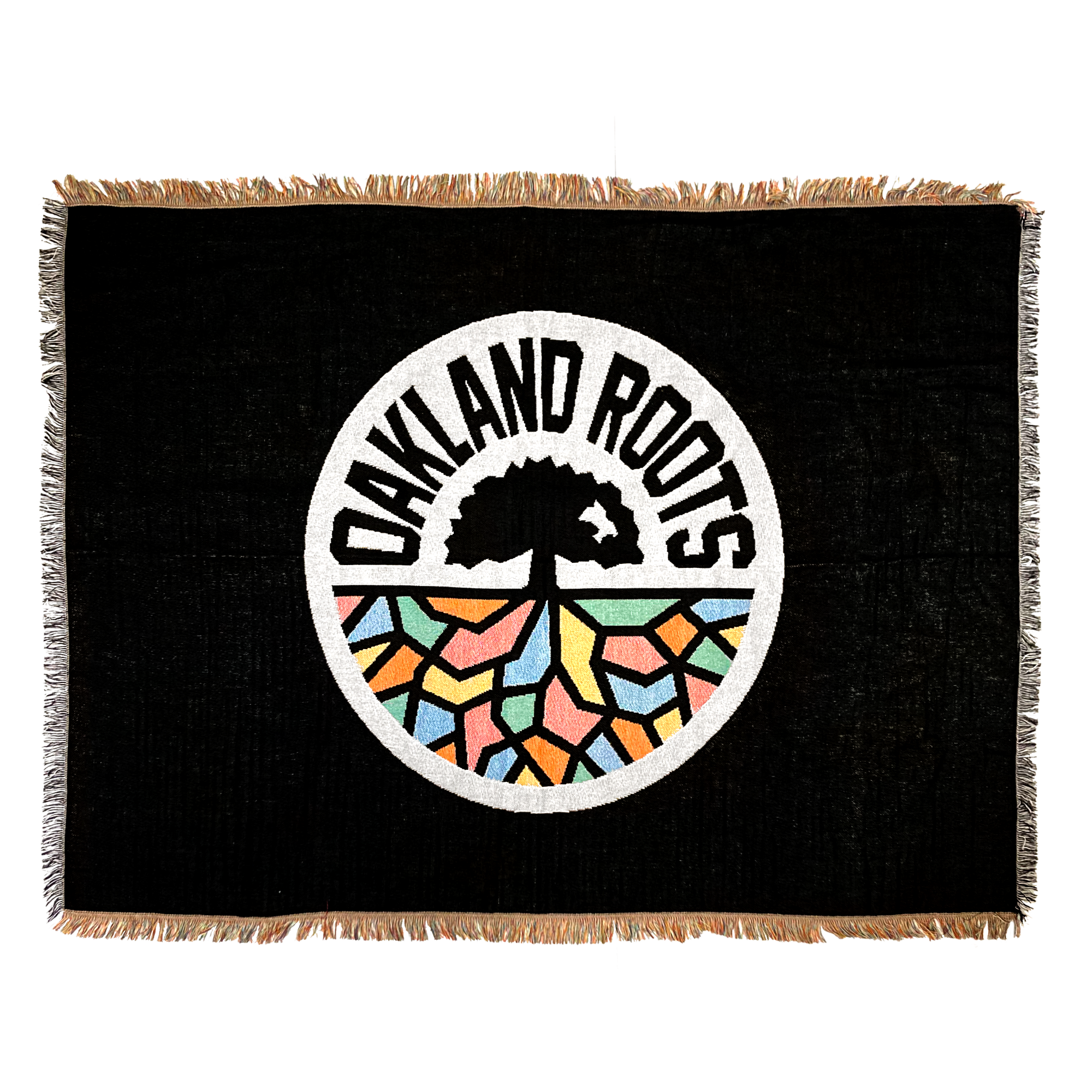 The Oakland Roots SC Throw is a rectangular black tapestry showcasing the Oakland Roots Sports Club logo in the center, which features a tree with sprawling roots above a vibrant, mosaic-like ground. The words "Oakland Roots" encircle the tree within a white-bordered circle. This socially conscious throw is accented with fringe along its edges.
