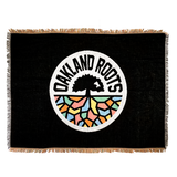 Black blanket with colored fringe and large full-color Oakland Roots SC circle logo in the center.