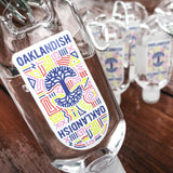 Close-up of multiples of travel-size hand sanitizer with Oaklandish logo and carabiner on wooden desk.