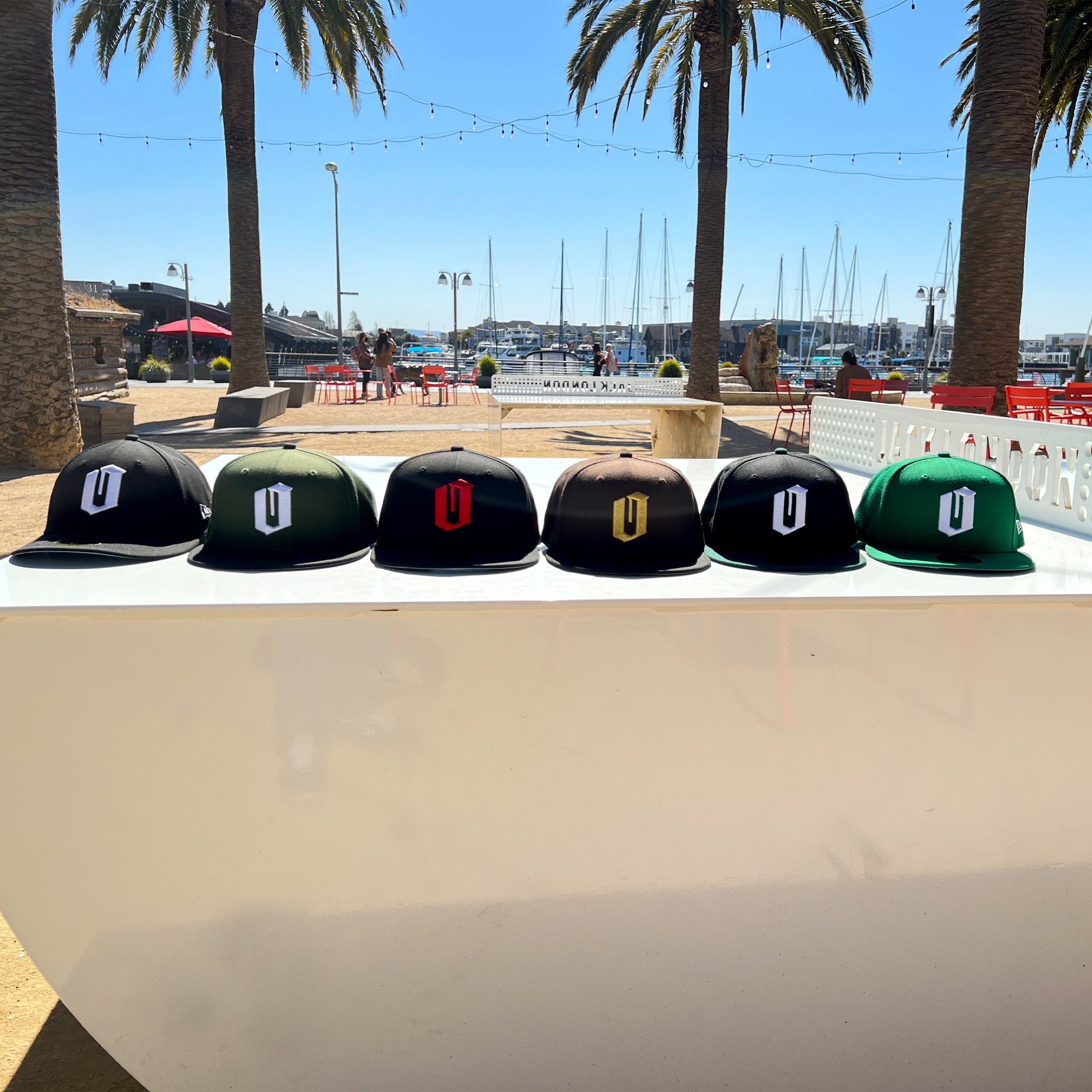 A row of six New Era Official 59FIFTY Fitted Caps, each featuring different designs, is showcased on a white surface outdoors. In the background, palm trees, string lights, red chairs, and a marina with boats docked under a clear blue sky can be seen. The Oaklandish cap stands out among the New Era collection.