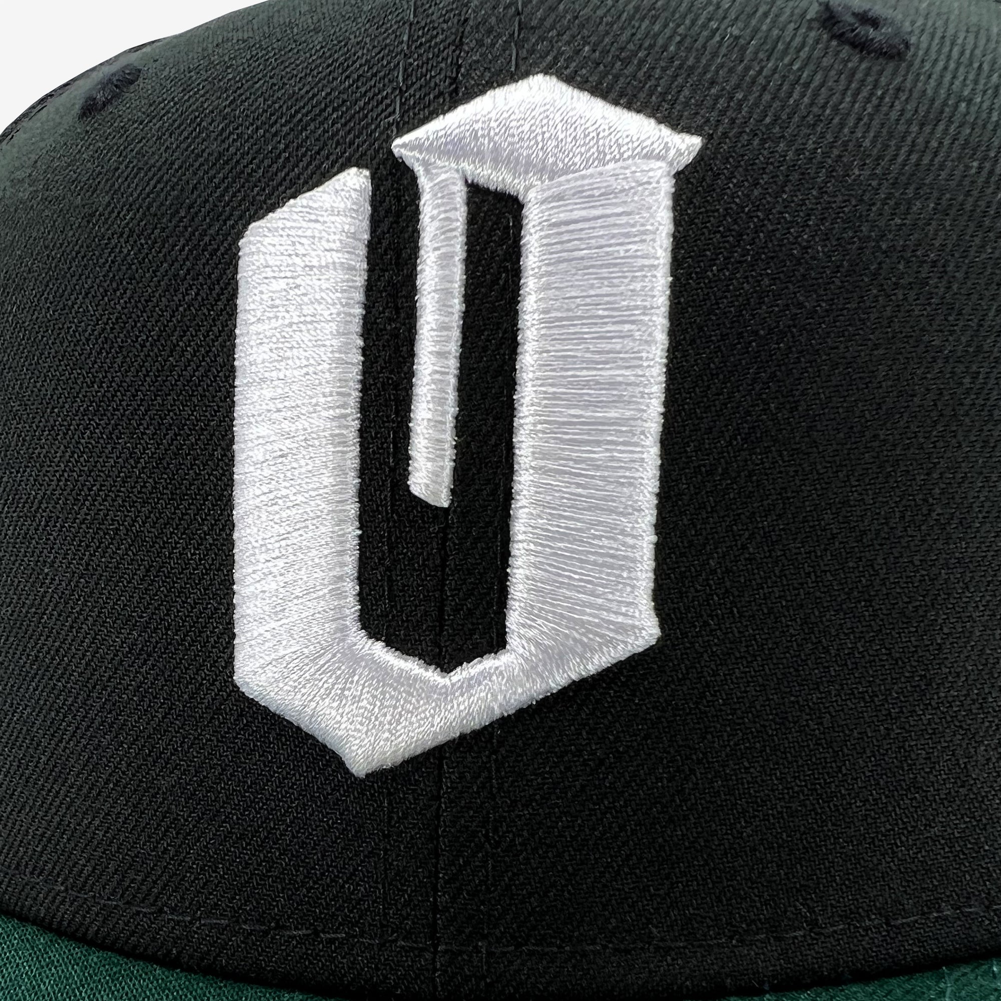 The Oaklandish New Era Official 59FIFTY Fitted Cap is a black baseball cap featuring a prominent, white embroidered "U" in a blocky, angular style at the center of the front panel. The stark contrast between the sharp-angled letter and the dark fabric enhances its distinct appearance. Precise stitching adds to its detailed design, capturing that signature Oaklandish flair.