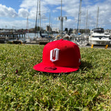 New Era 9FIFTY red cap with white embroidered O siting on grass in Oakland park with boats in the background.