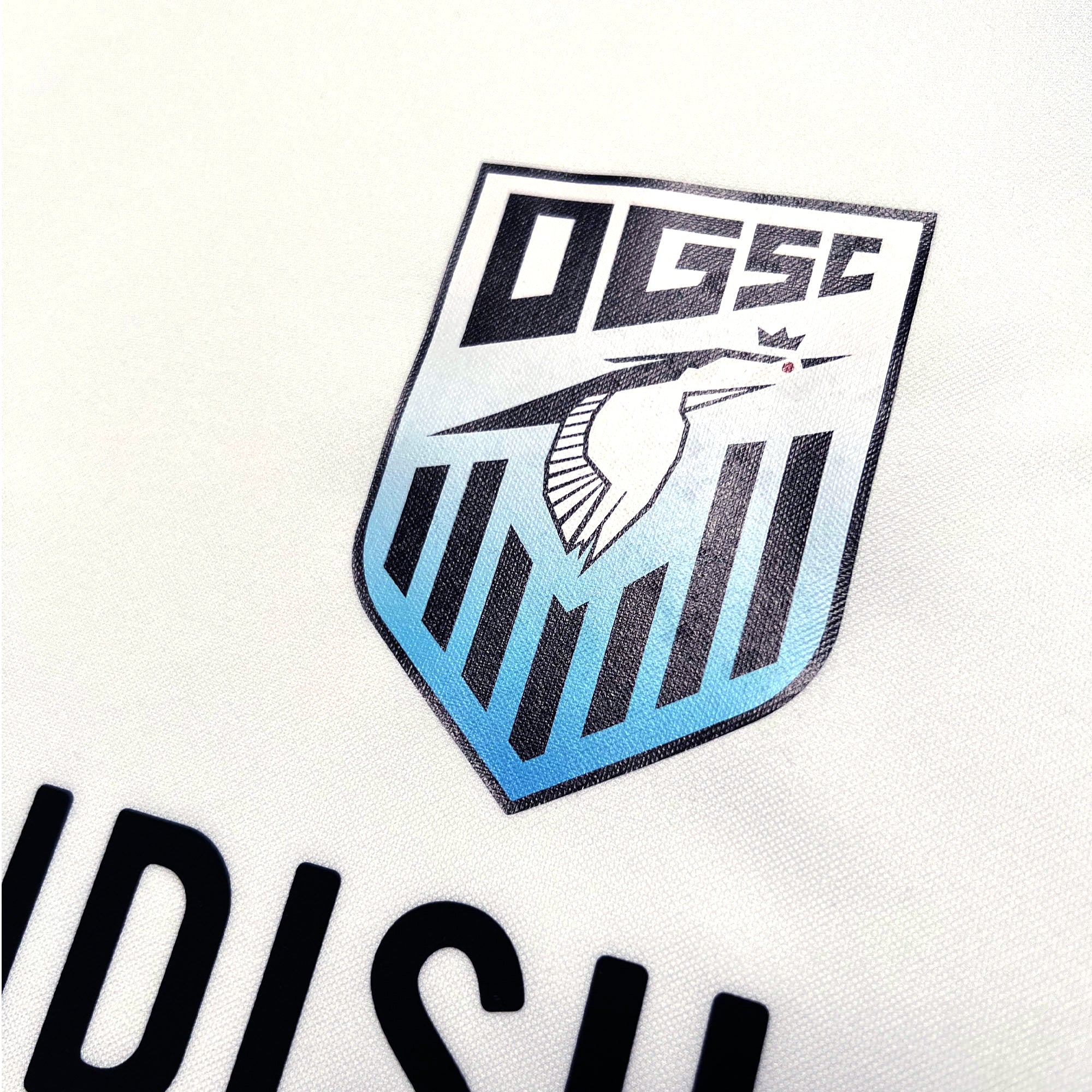 Detailed close-up of Oakland Genesis heron logo on a white soccer jersey. 