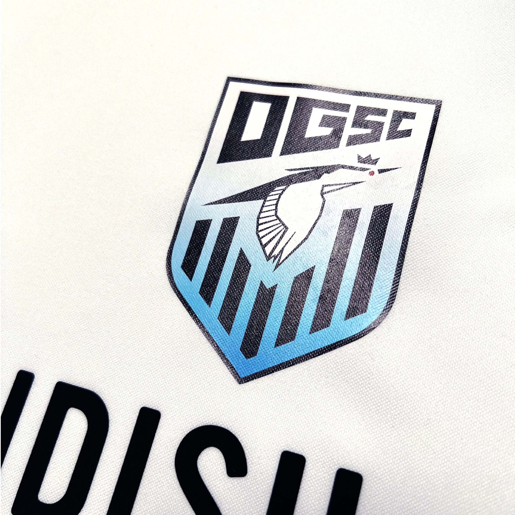 Close-up image of the Oakland Genesis Home White jersey by Partner. The youth soccer team's logo features a stylized kingfisher with a crown on its head, placed inside a shield adorned with vertical stripes. The letters "OGSC," symbolizing Oakland Genesis, are prominently displayed at the top of the shield.