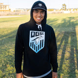 A man standing on a soccer field wearing a black pullover hoodie with an Oakland Genesis soccer league heron logo on chest.