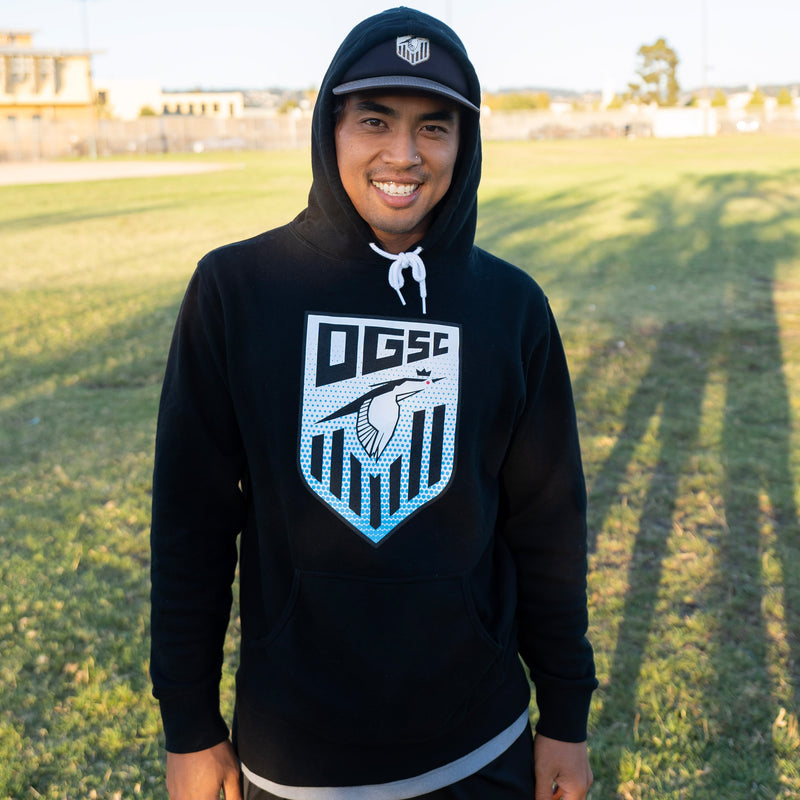 A man standing on a soccer field wearing a black pullover hoodie with an Oakland Genesis soccer league heron logo on chest.