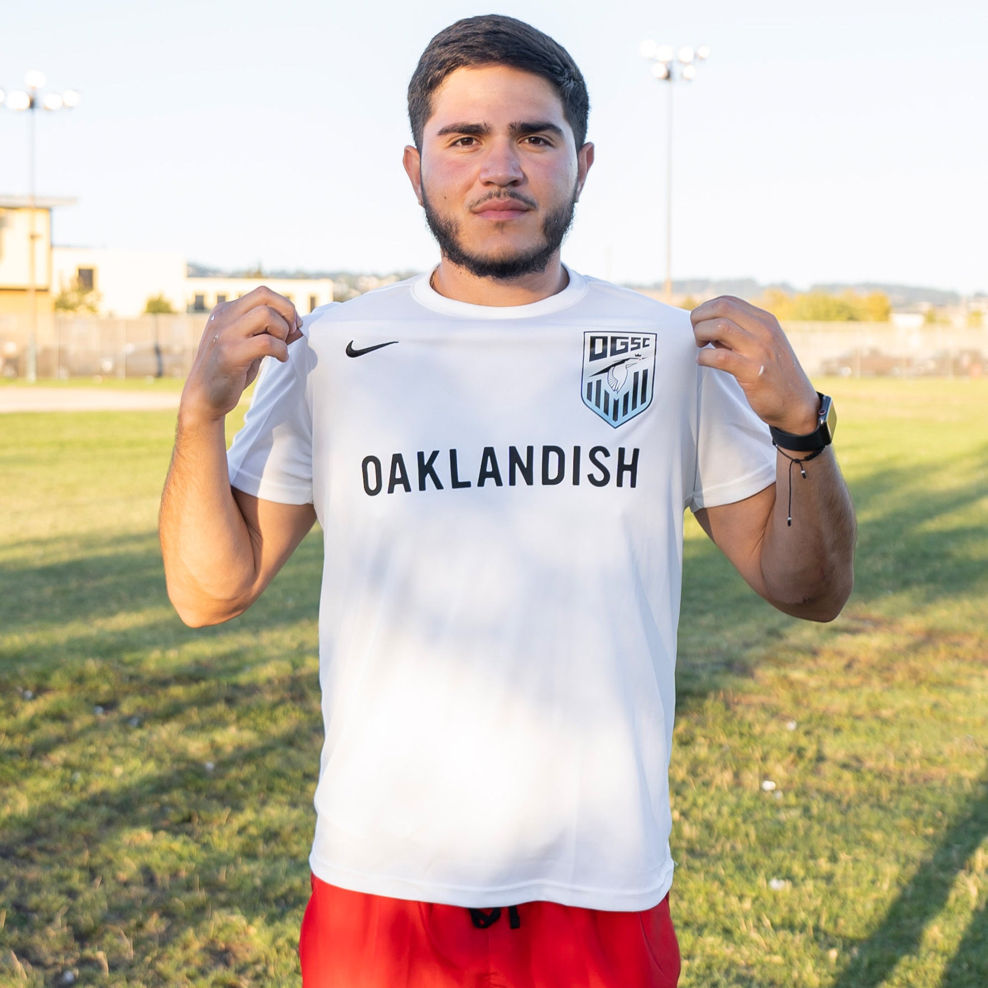 A man with short brown hair and a beard stands outdoors on a grassy field. He is wearing the Oakland Genesis Home White soccer jersey by Partner, featuring "Oaklandish" across the front and an Oakland Genesis logo on the left. The man is holding the sides of his jersey and looking directly at the camera, while sporting a black wristwatch.